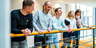 Vijf medewerkers naast elkaar leunend tegen railing